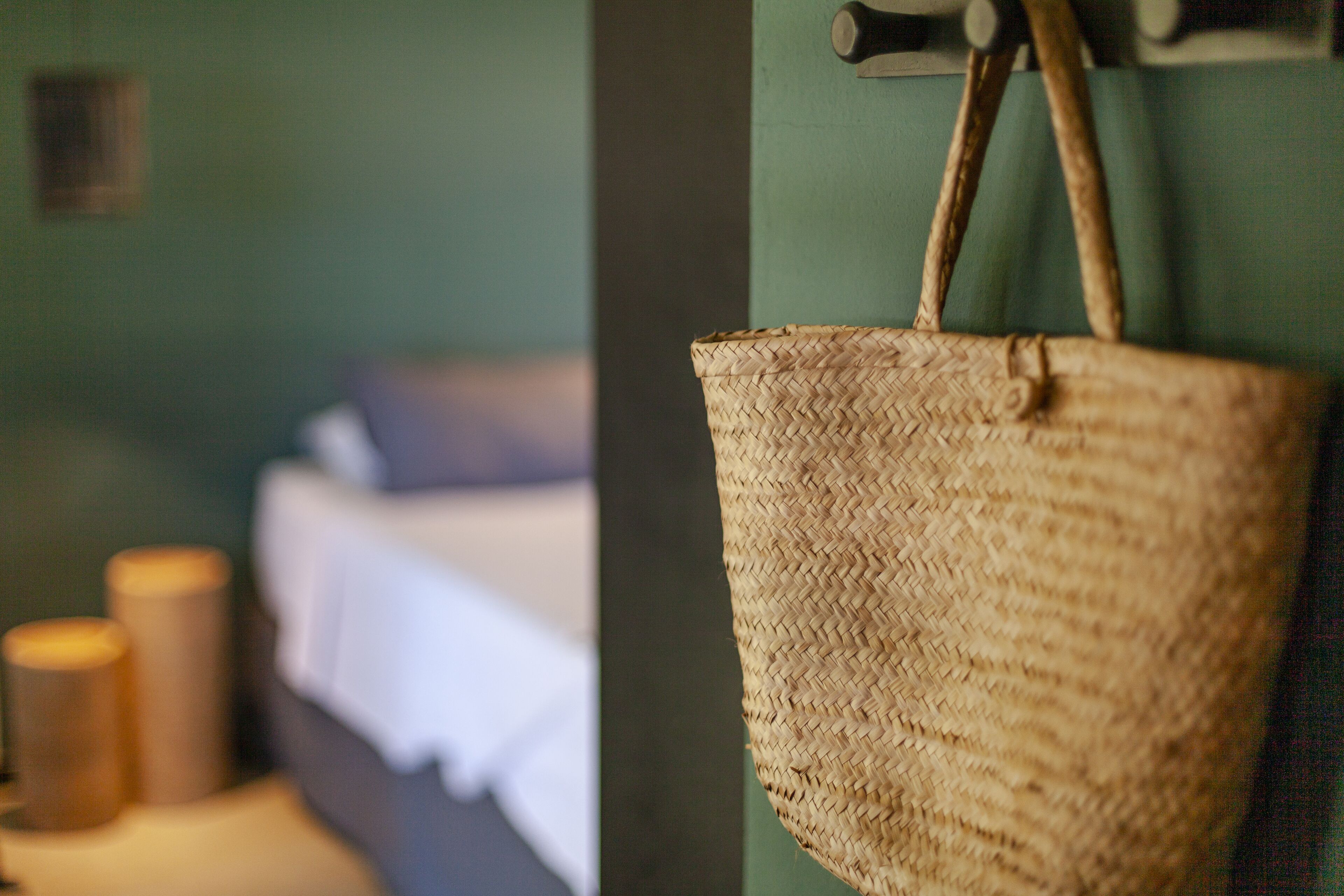 Una cesta de mimbre colgada en una pared, una cama con sábana blanca y dos velas sobre una mesita de noche.