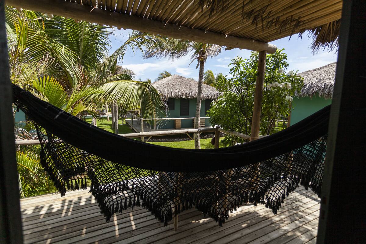 A hammock on a wooden deck with a view of a thatched-roof building and palm trees.