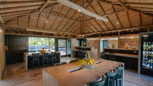 Un espacio interior amplio con una gran mesa de madera rodeada de sillas, una zona de bar con un refrigerador lleno de botellas y vistas a zonas verdes.