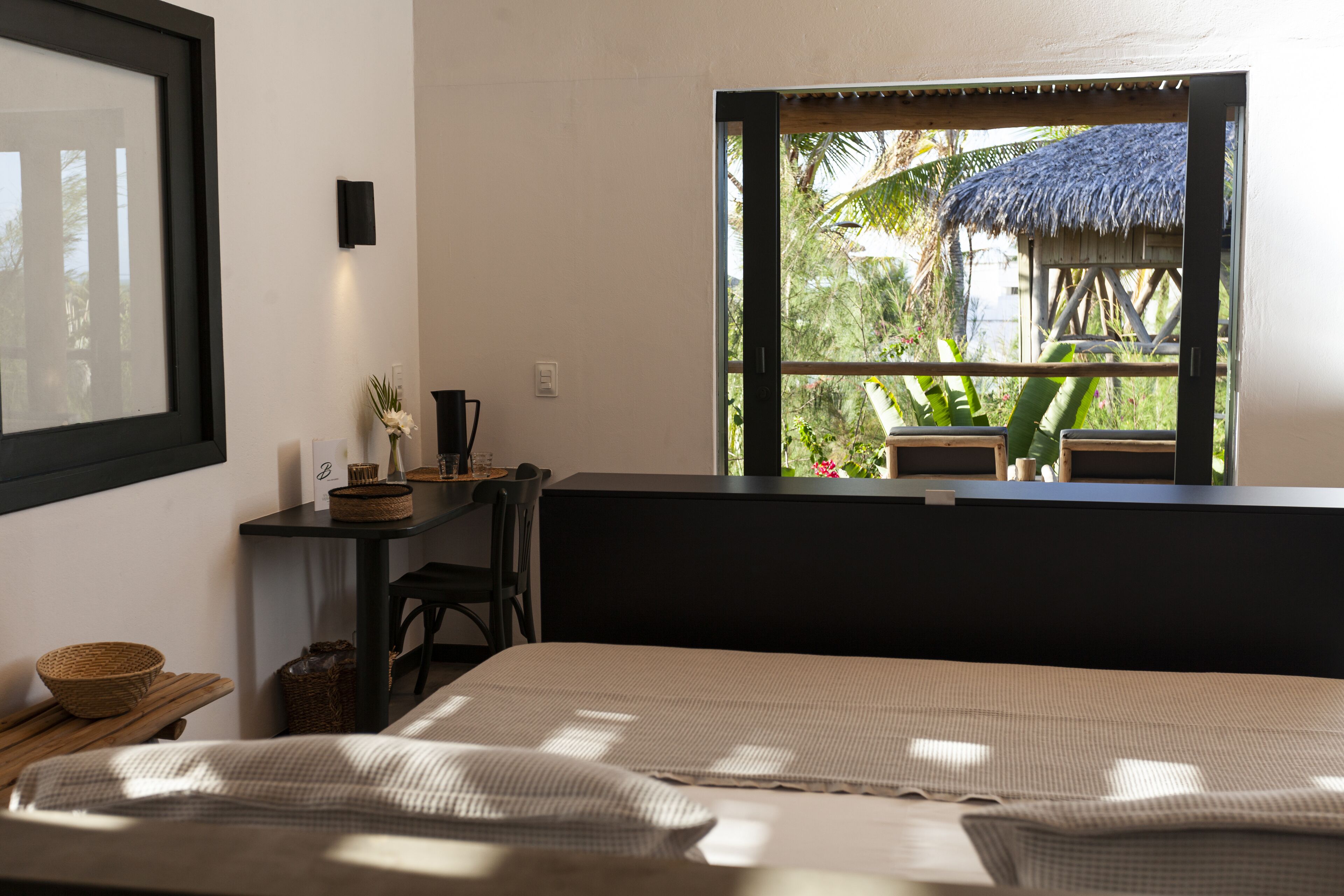 A bedroom with a bed, a desk, and a window overlooking a thatched-roof structure.