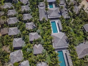 Vista aérea de un resort con cabañas de techo de paja dispuestas en filas, rodeado de exuberante vegetación y con piscinas centrales.