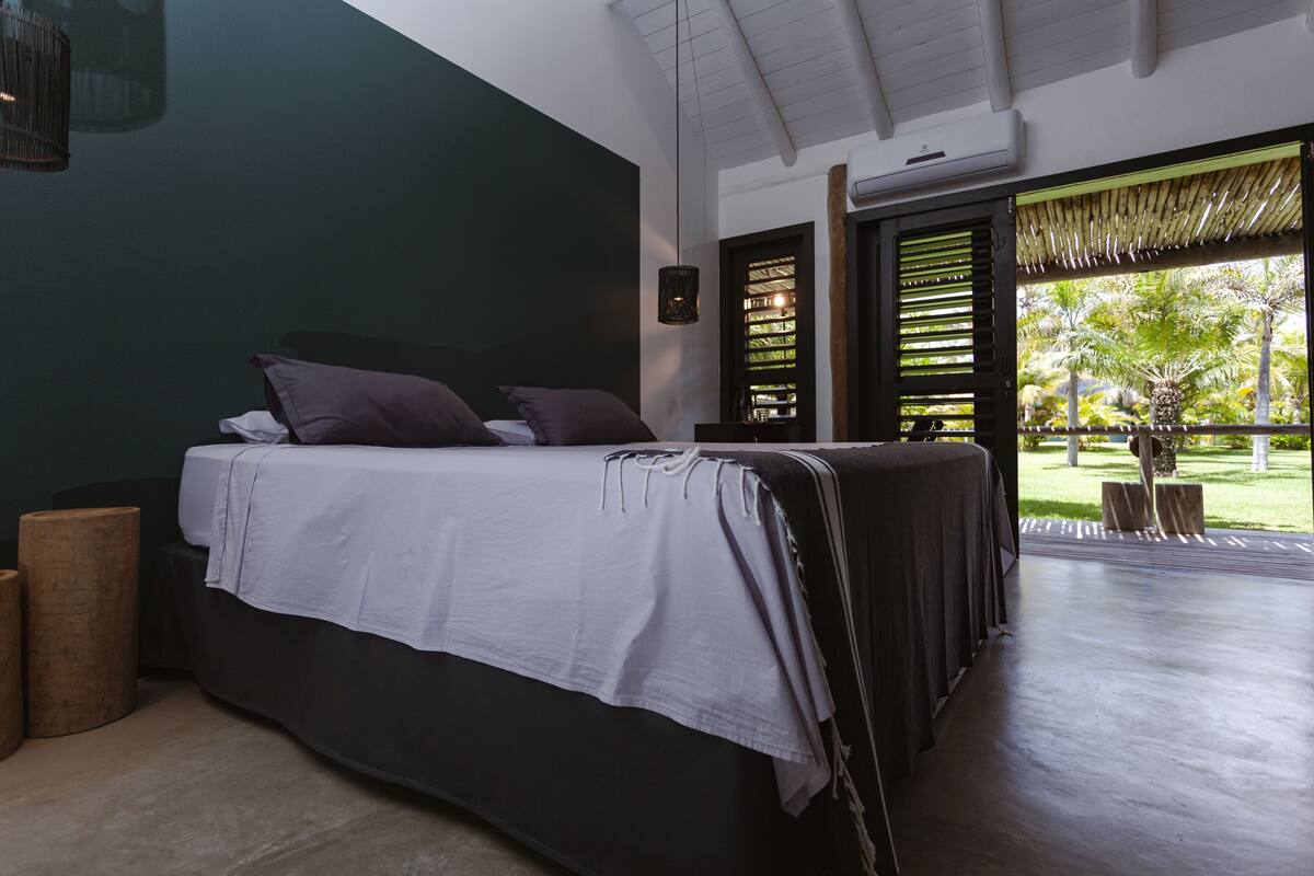 A bedroom with a bed, a nightstand, a window with blinds, and a view of greenery outside.