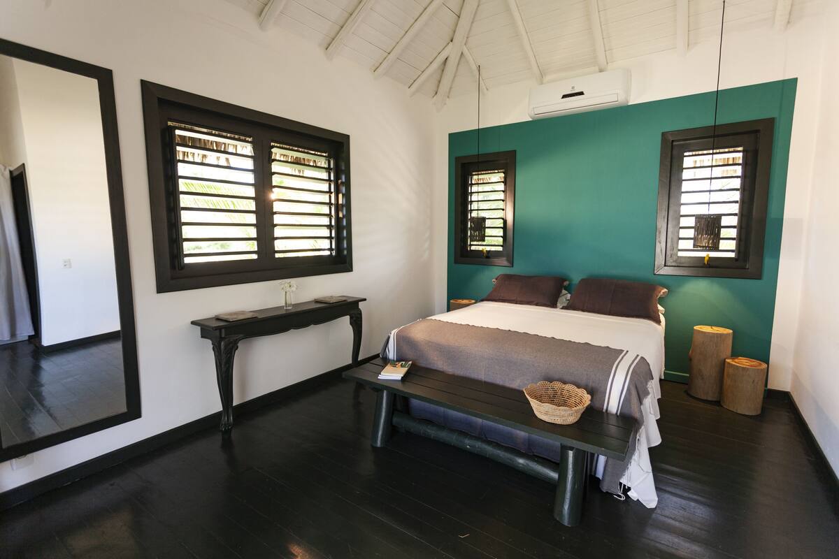A bedroom with a bed, a bedside table, a mirror, and two windows with blinds.