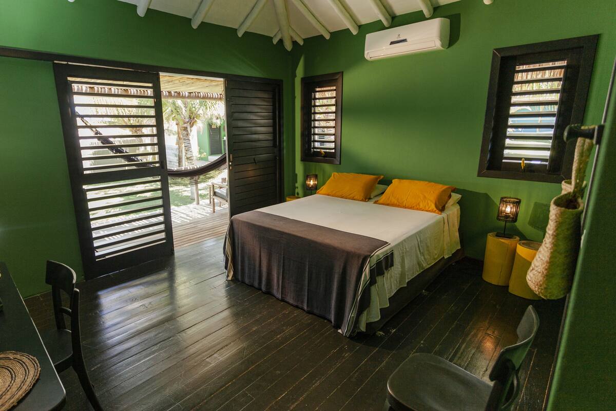 A room with a bed, a chair, and a table. There is a window with blinds and a door leading to an outdoor area.