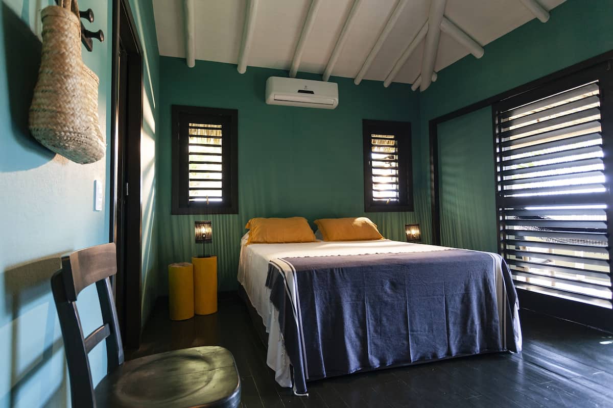 A bedroom with a bed, a chair, a hanging bag, and two windows with blinds.