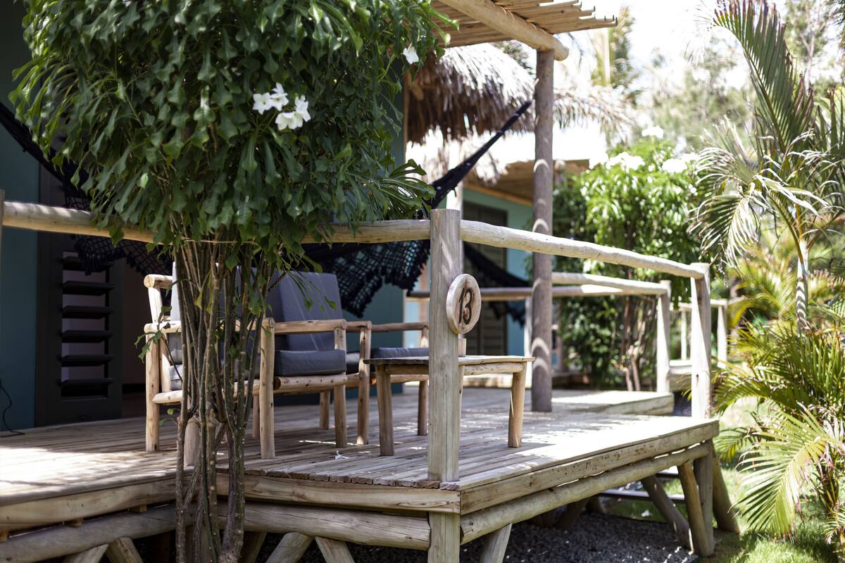 A wooden deck with a thatched roof, surrounded by lush greenery and a palm tree.