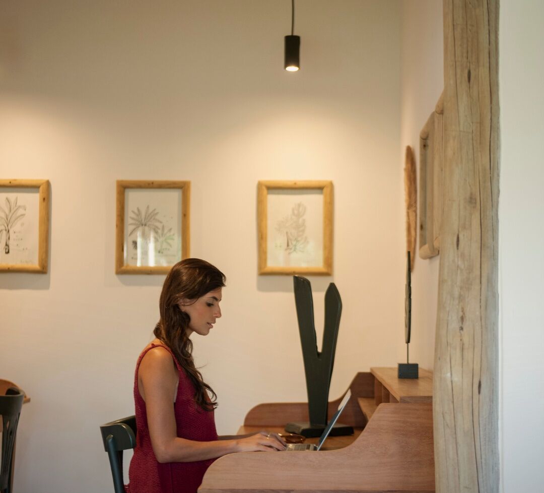 Una mujer sentada a una mesa de madera con un ordenador portátil, en una habitación con ilustraciones botánicas enmarcadas en la pared.