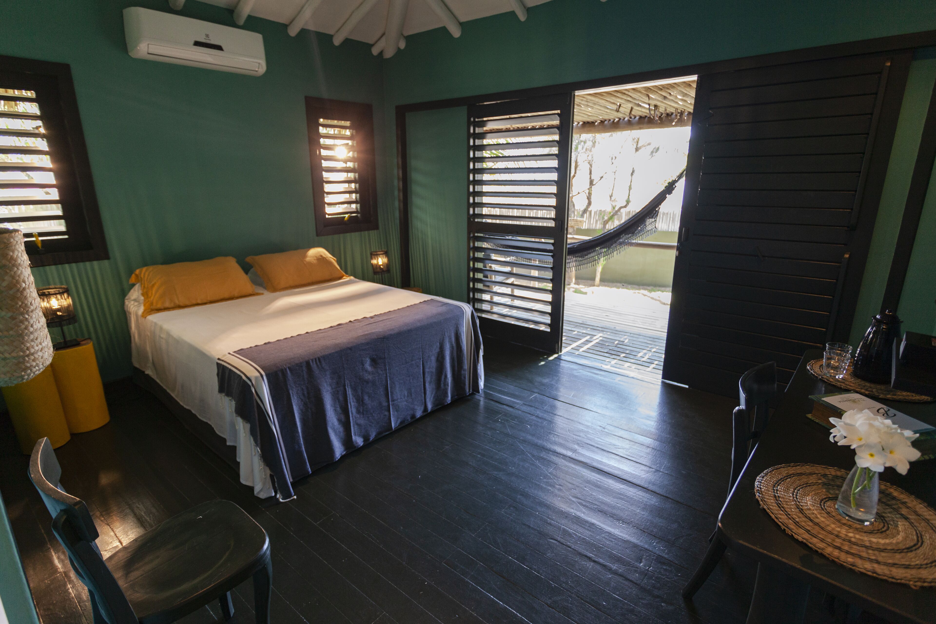 A room with a bed, a chair, a table with a vase of flowers, and a hammock outside.