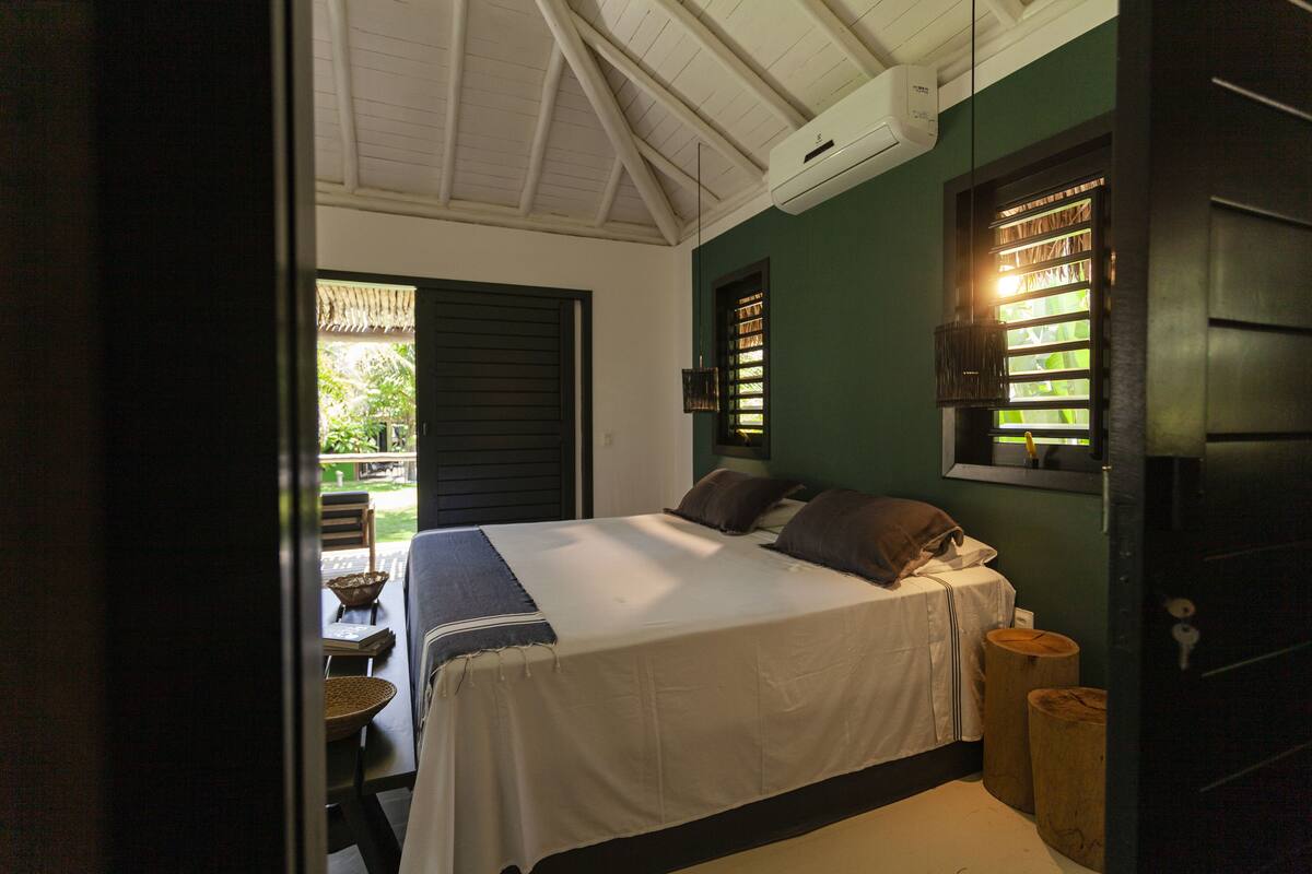 A bedroom with a bed, a window with blinds, a wooden stool, and a table with chairs.