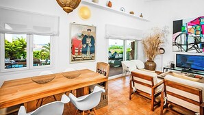 A spacious living area with a wooden dining table, white chairs, a sofa, and a television. There is a large painting on the wall and a decorative hanging light fixture.