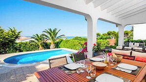 An outdoor dining area with a pool, wooden table, and chairs, overlooking a pool and greenery.