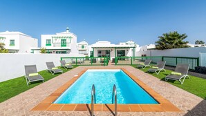 A swimming pool with a tiled deck and lounge chairs, surrounded by white buildings with green accents and a clear blue sky. - Villa Pargo - Three Bedroom Villa, Sleeps 6 (Playa Blanca)
