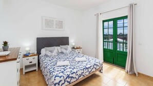 Een slaapkamer met een bed, nachtkastjes, een bureau, een potplant en een groene deur met uitzicht.
