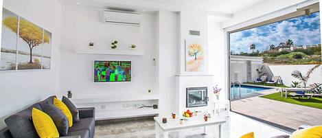 A modern living room with a flat-screen TV, a white coffee table, and a grey sofa with yellow cushions. There is a large window with a view of a pool and greenery.