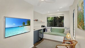A compact room with a large TV displaying a beach scene, a small kitchenette, a window with a view of greenery, and a cozy seating area with cushions. - Hibiscus Hideaway Valla Beach 4BR home w/ two verandahs (Valla Beach)