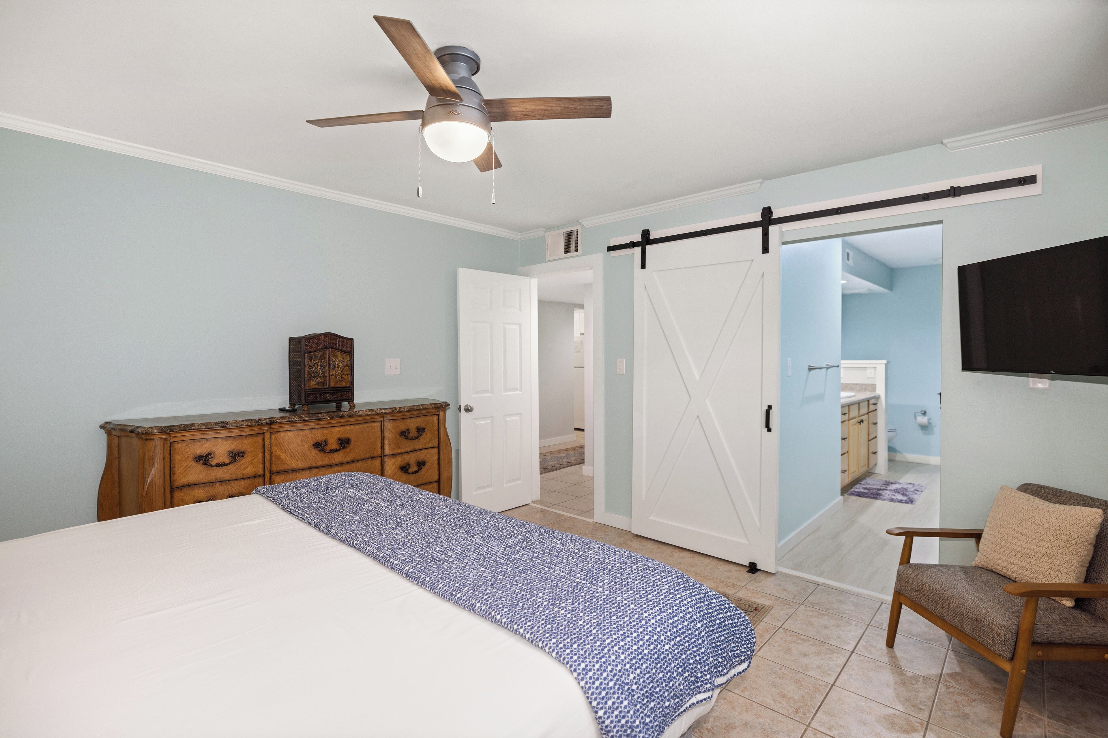 A bedroom with a bed, a dresser, a chair, a ceiling fan, and a sliding door leading to a bathroom.