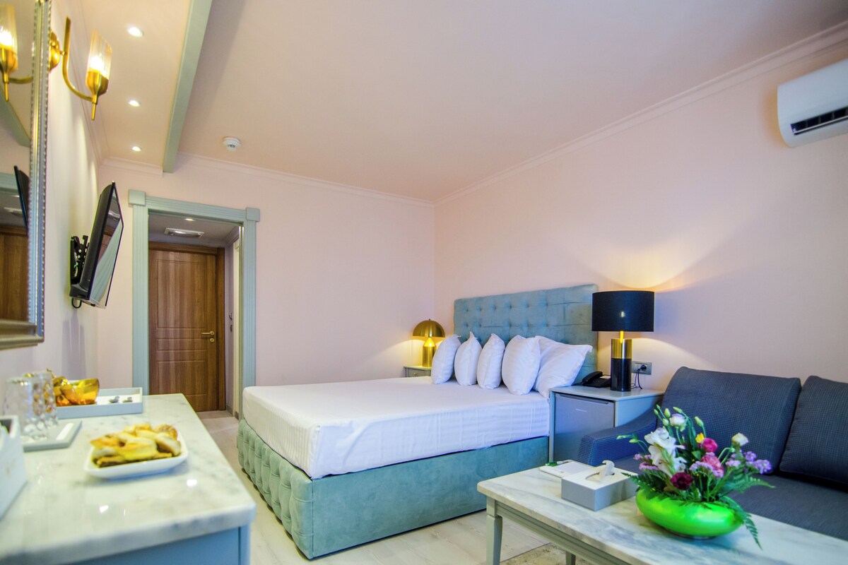 A hotel room with a bed, a sofa, a bedside table, a lamp, and a flower vase.