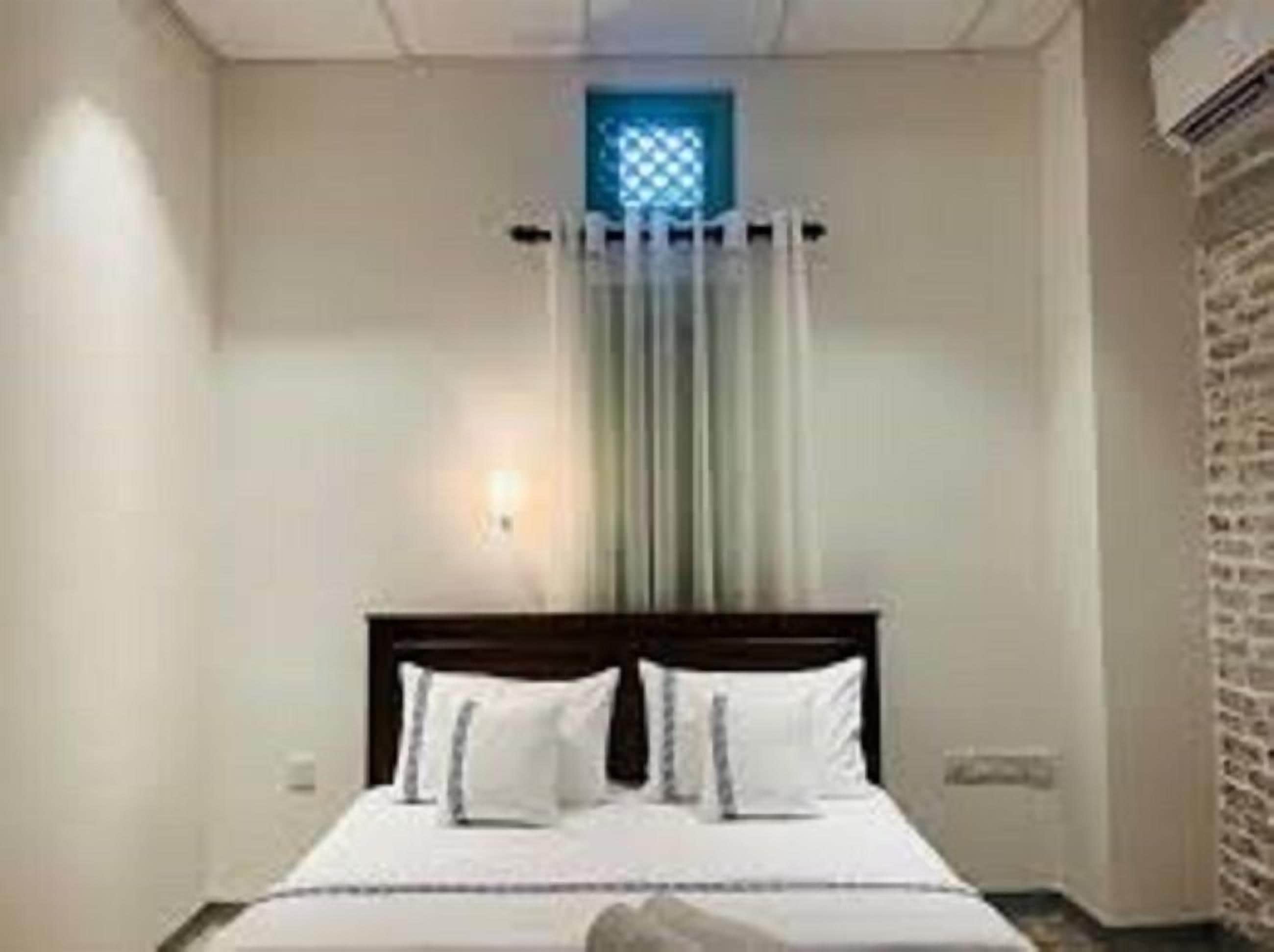 A hotel room with a bed, a window with a blue light, and a brick wall.