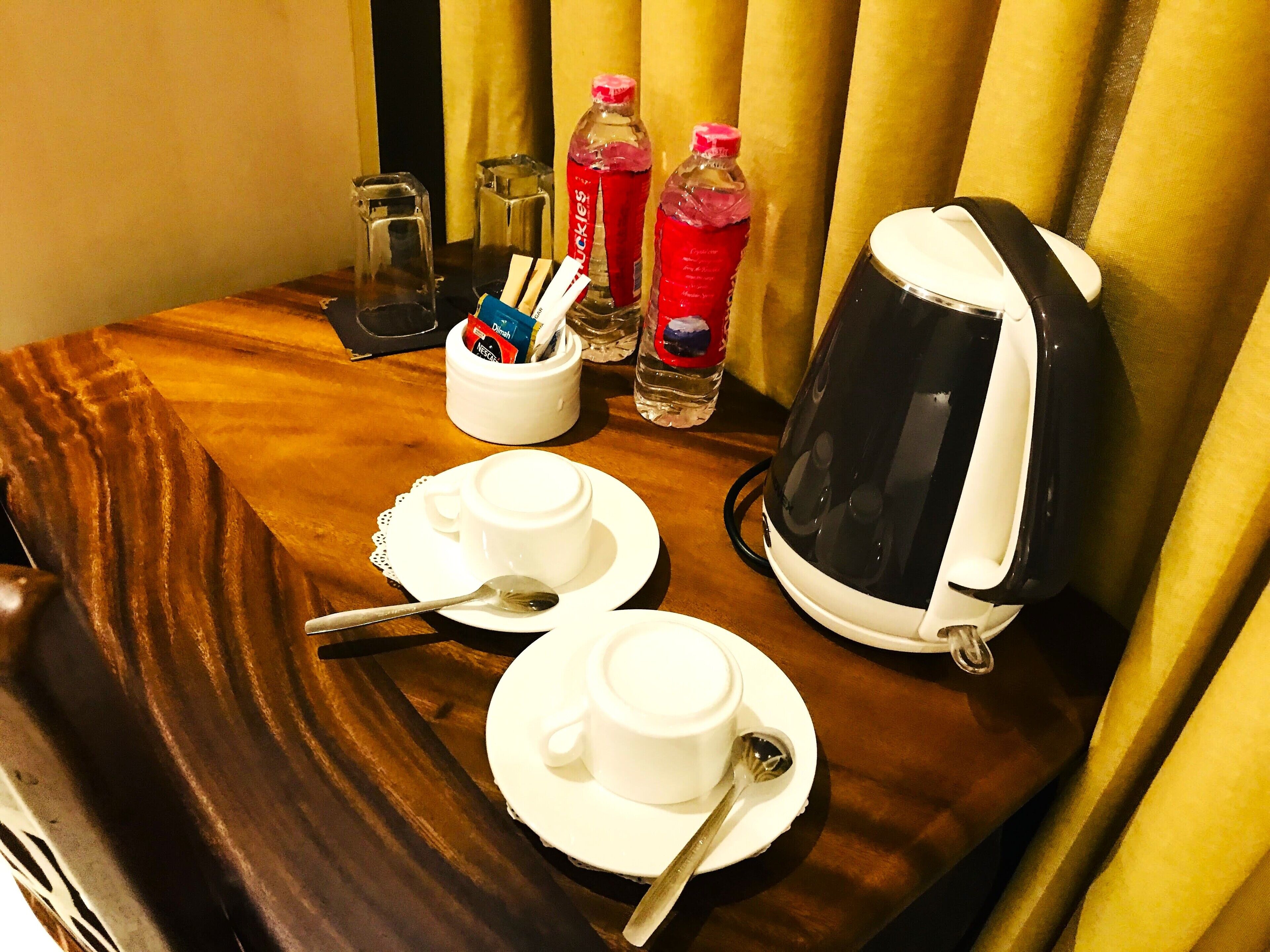 Una mesa de habitación de hotel con dos tazas blancas, cucharas, una tetera y dos botellas de agua.