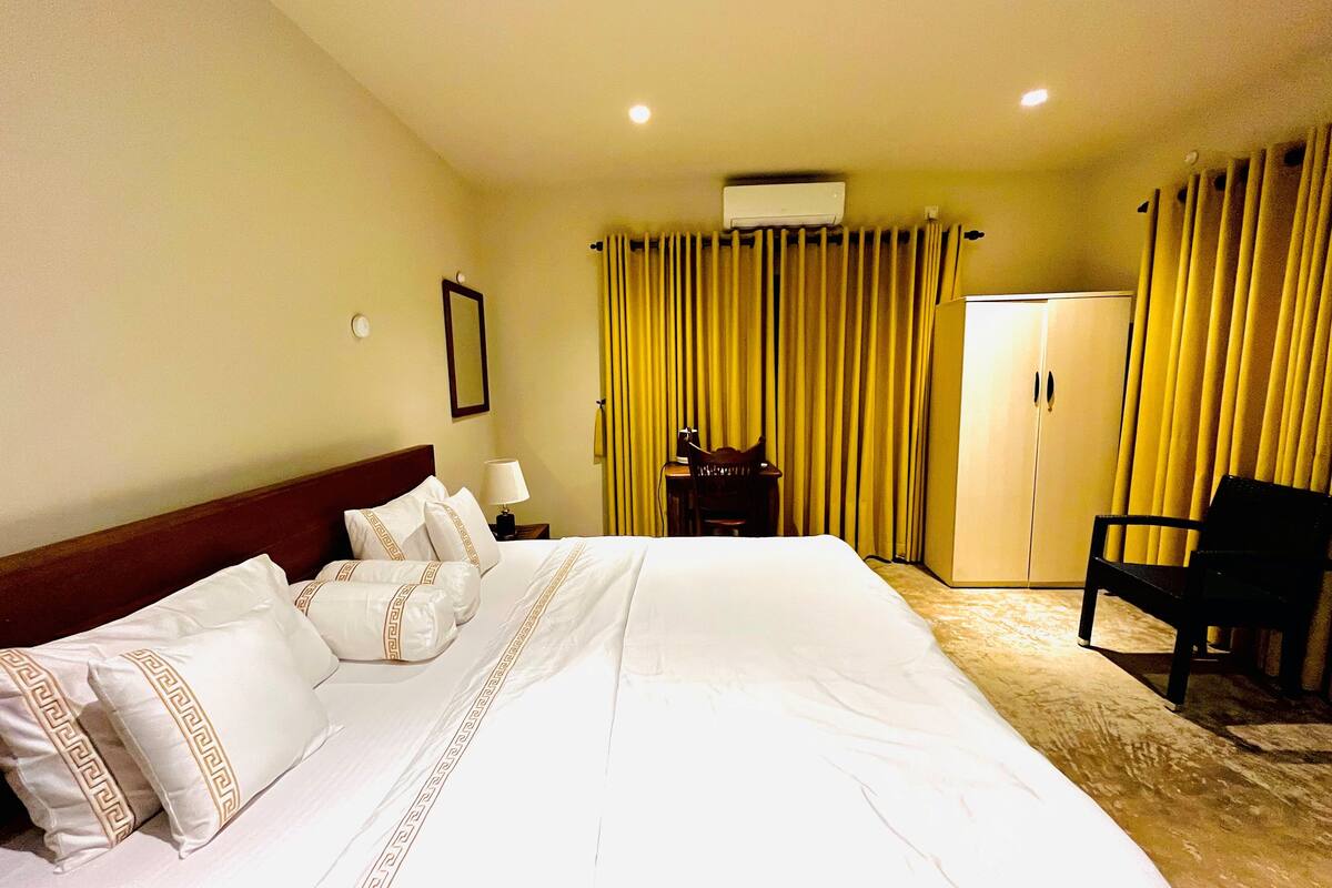 A hotel room with a large bed, a desk with a chair, a wardrobe, and an air conditioning unit on the wall.