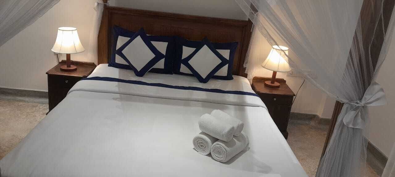 A neatly made bed with a white duvet, two pillows, and rolled towels. There are two bedside tables with lamps and a canopy over the bed.