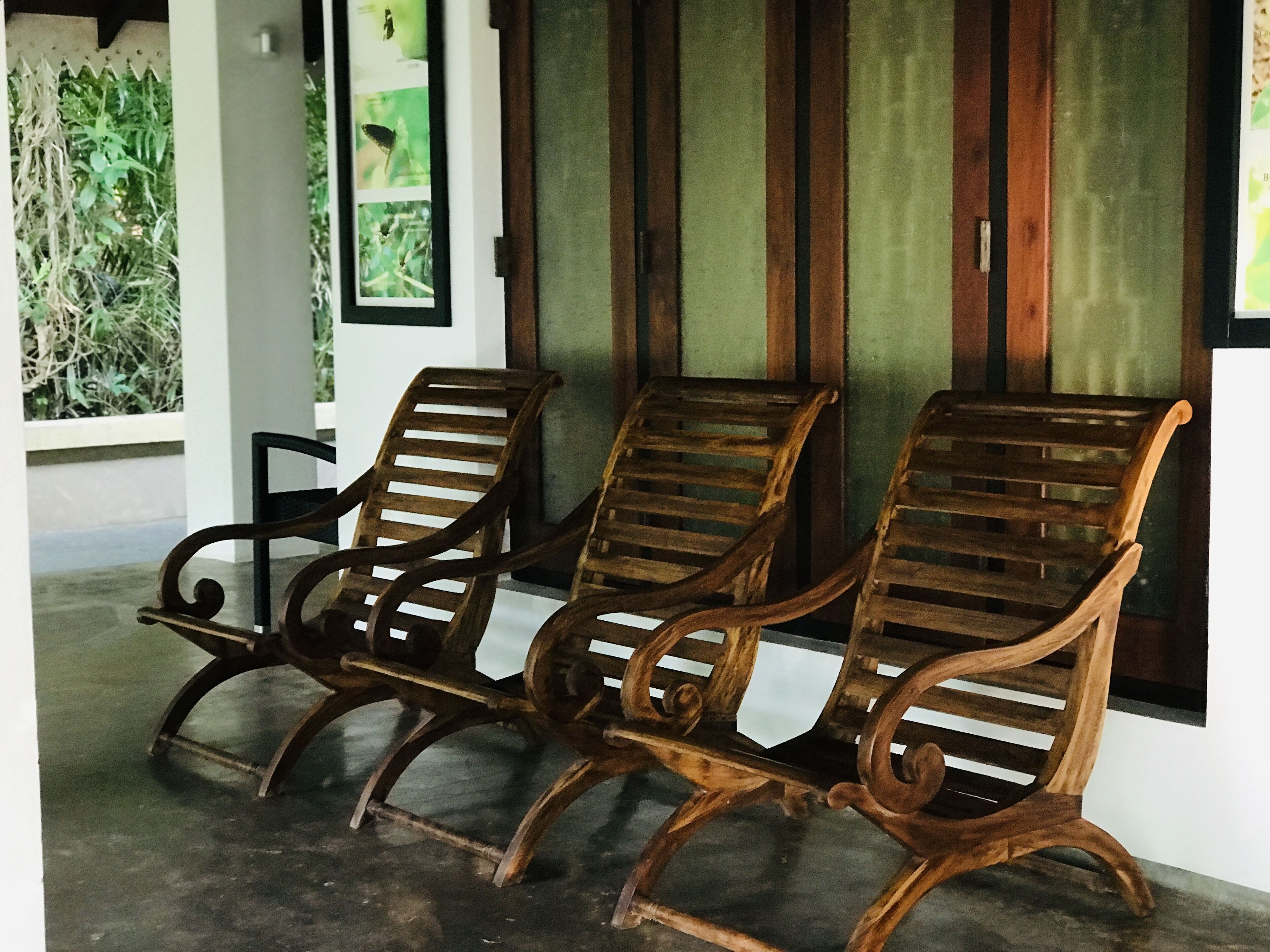 Three wooden lounge chairs with metal frames, positioned in front of a window with vertical blinds.