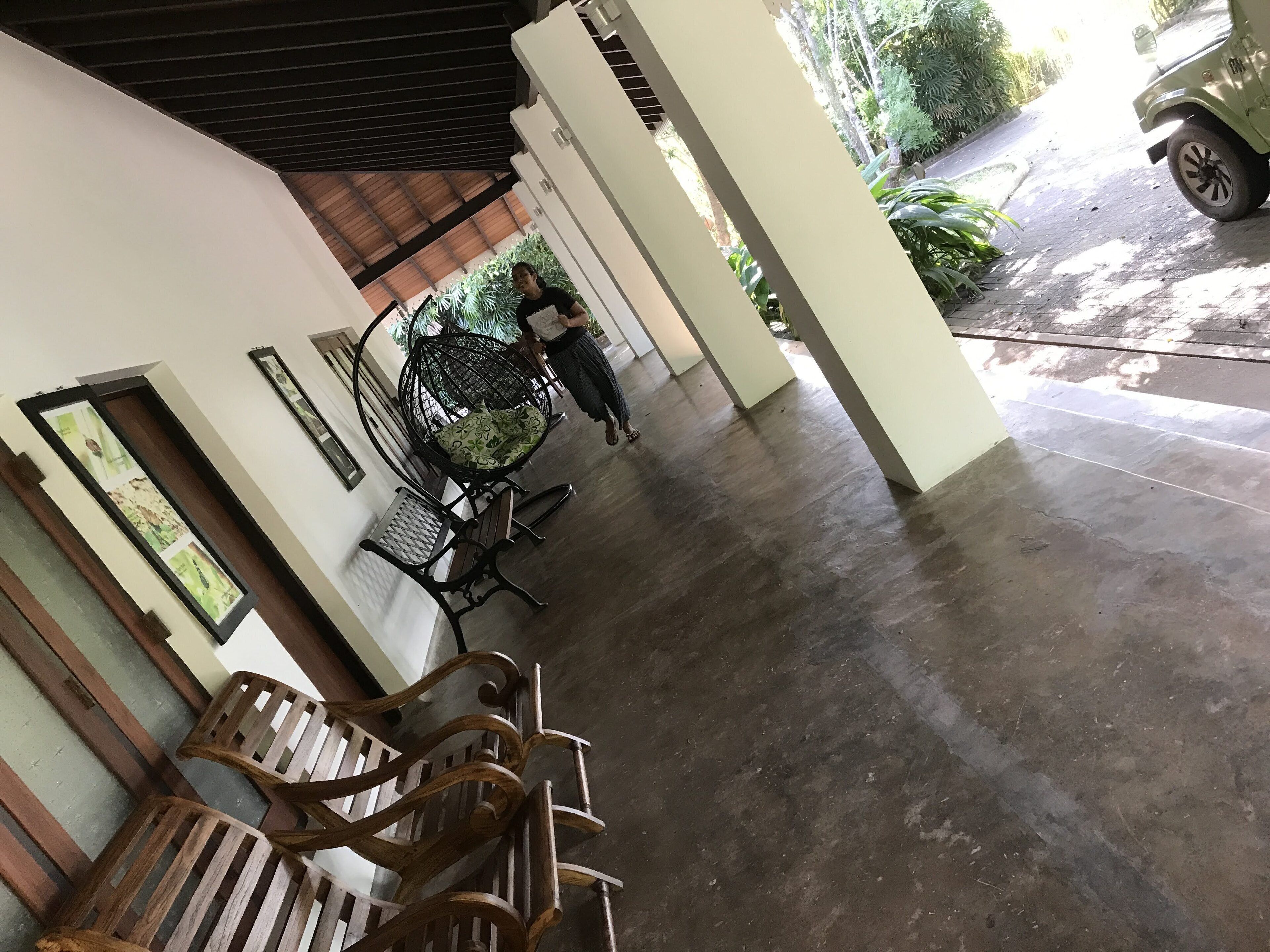 Un espacio al aire libre cubierto con piso de concreto, sillas de madera, una silla colgante y un vehículo estacionado fuera.
