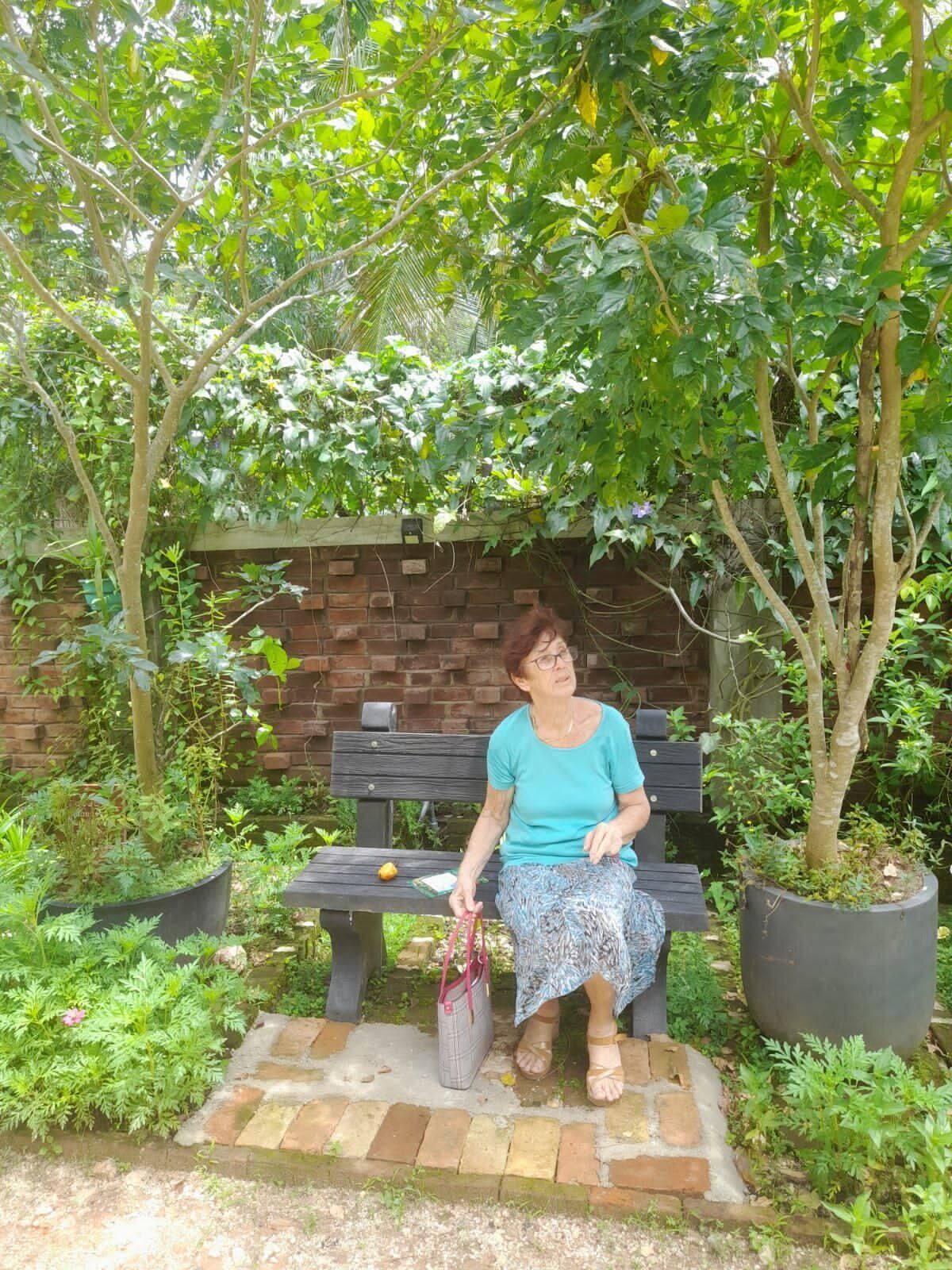 Una dona asseguda en un banc d'un jardí amb mur de maó i plantes en testos.