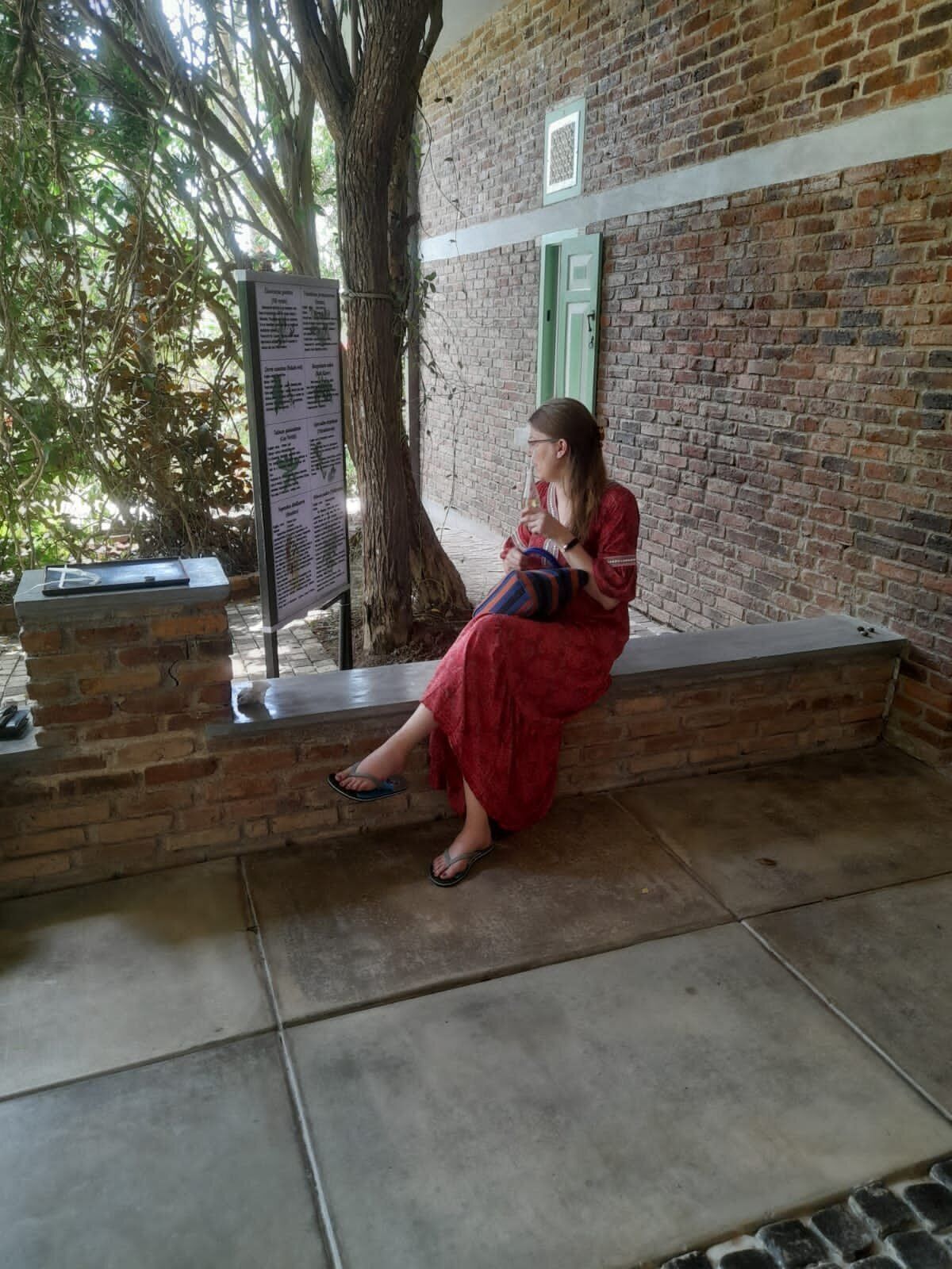 A person sitting on a bench in a covered outdoor area with a brick wall and a tree.