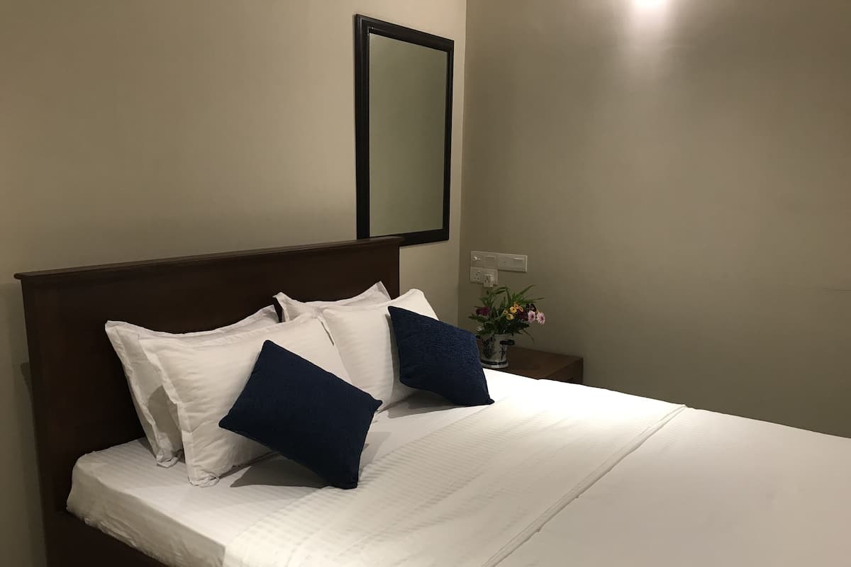 A neatly made bed with white and dark blue pillows, a wooden headboard, a nightstand with a vase of flowers, and a wall-mounted mirror.