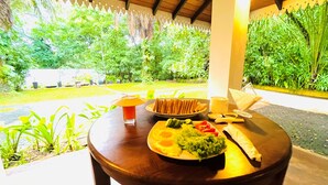 A table with food and drinks, surrounded by lush greenery and a covered area with a wooden ceiling. - Ceylon Kingsmen Garden Hotel (Negombo)