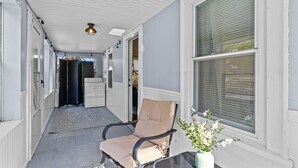 Un couloir avec un fauteuil beige, une petite table sur laquelle est posé un vase de fleurs et une fenêtre avec des stores.