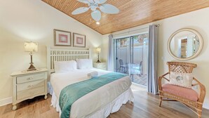 Ein Schlafzimmer mit einem Bett, Nachttischen, einem Deckenventilator, einem Fenster mit Vorhängen und einem Korbstuhl.