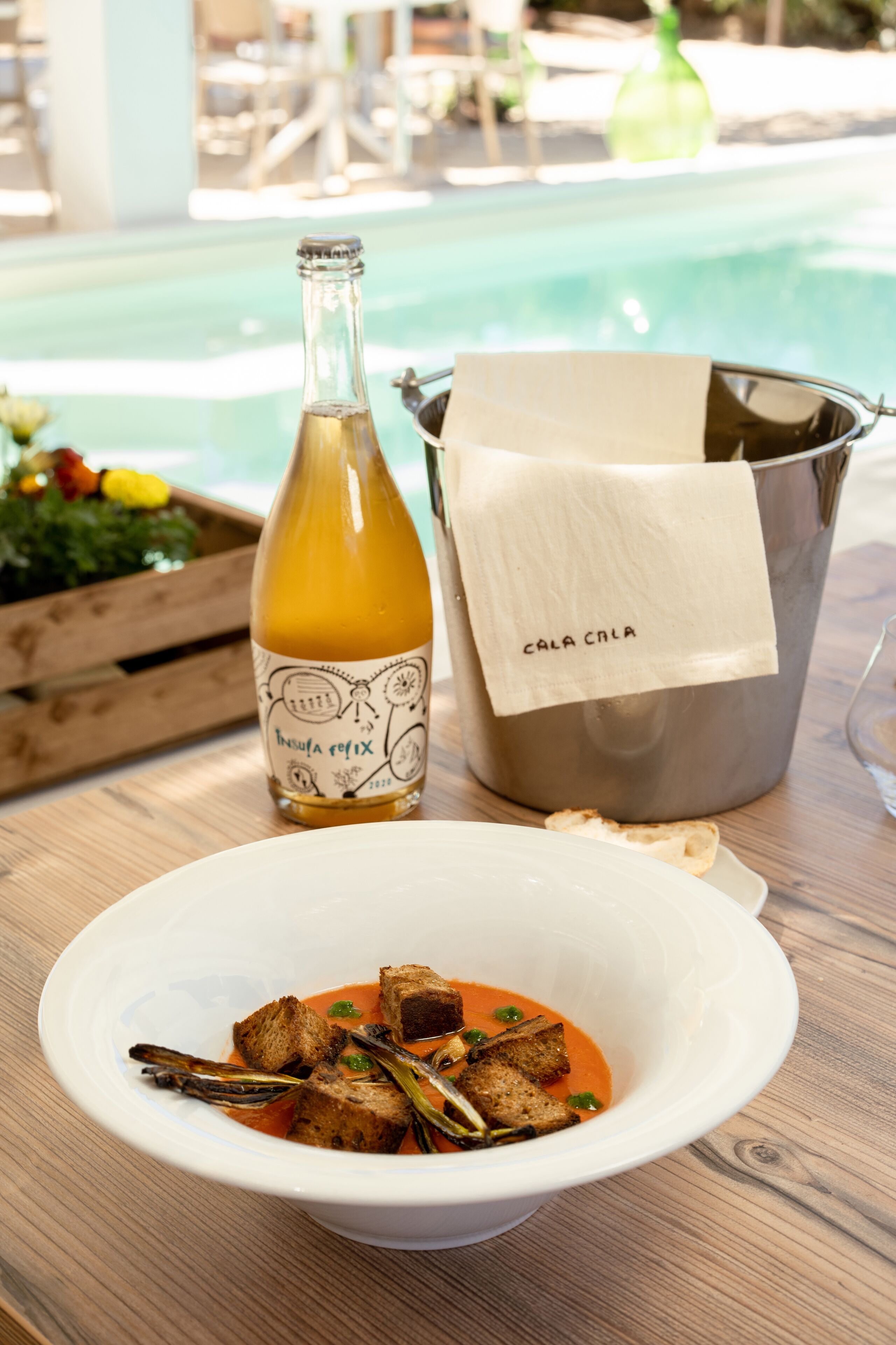 A dining table with a bowl of soup, a bottle of wine, and a bucket labeled "CALA CALA".