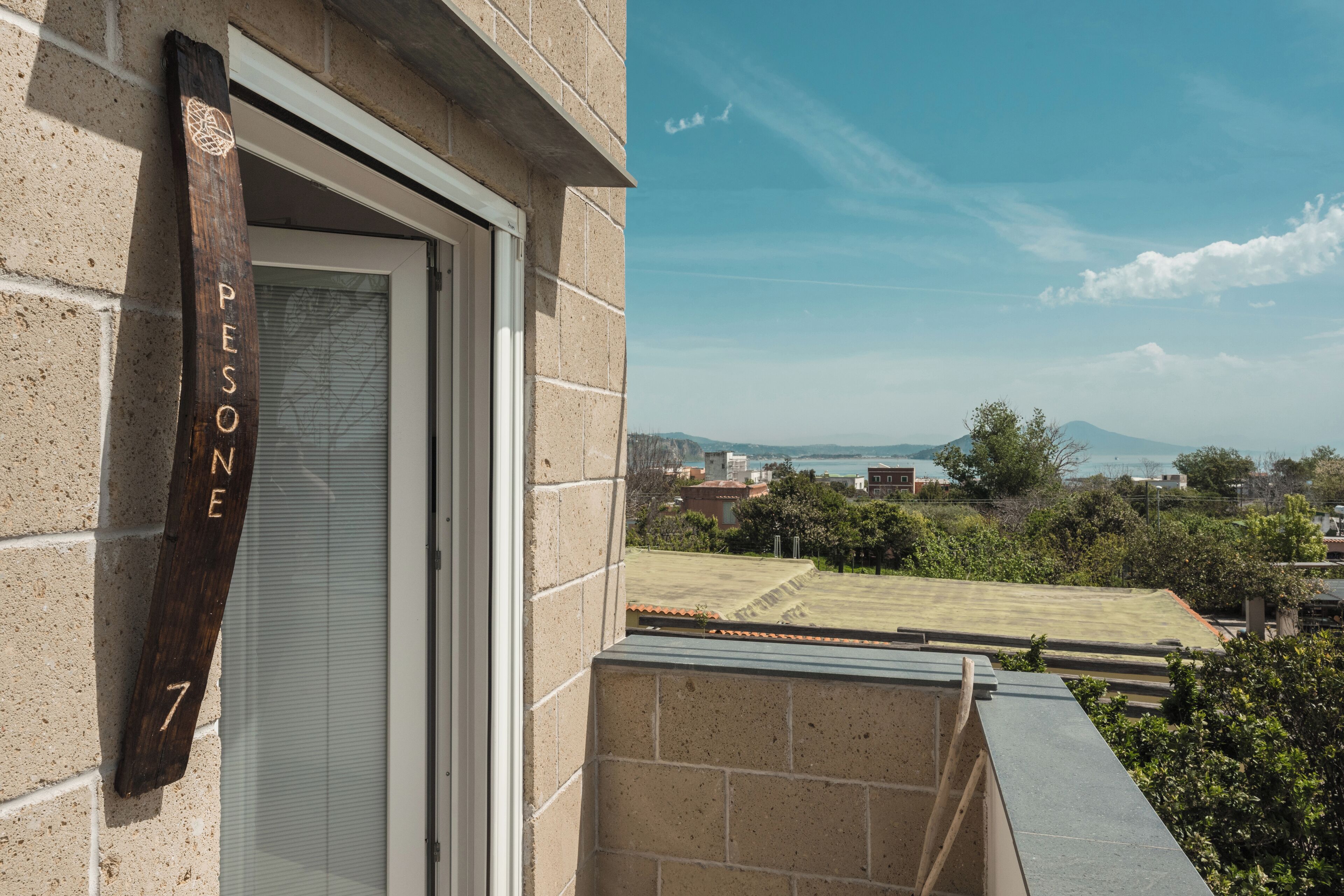 A wooden sign with "PESONE 7" on a building facade, a balcony with a view of a green field and distant mountains.