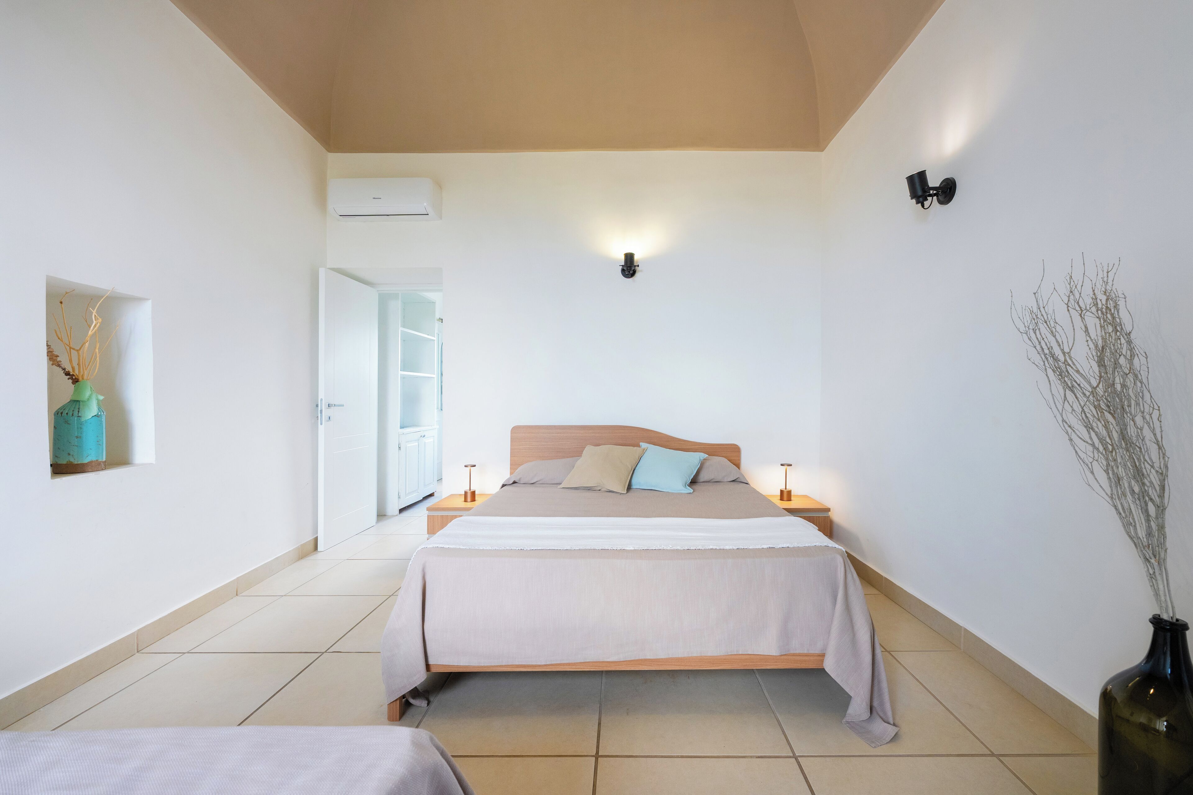 A bedroom with a bed, bedside tables, a nightstand, a wall-mounted light, and a vase with branches.