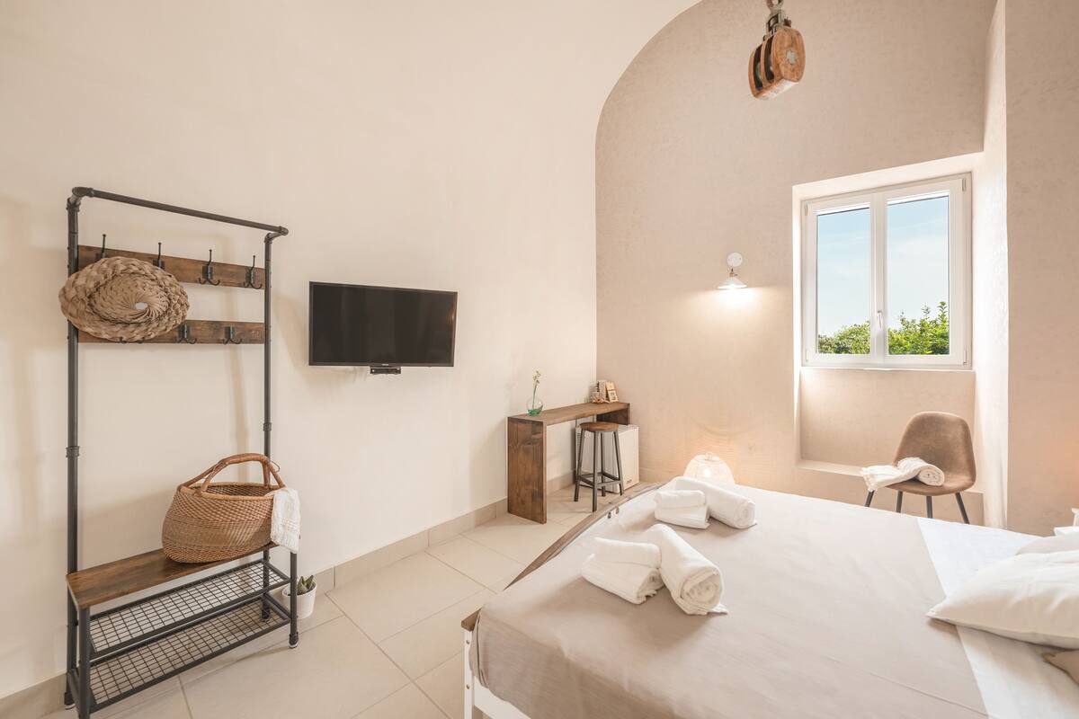 A bedroom with a bed, a TV, a desk, a chair, a window, and a shelf with a basket and towel.