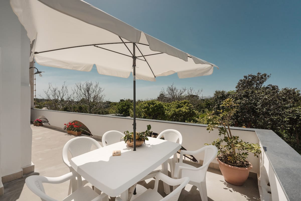 A rooftop terrace with a white table and chairs, a large umbrella, and potted plants.