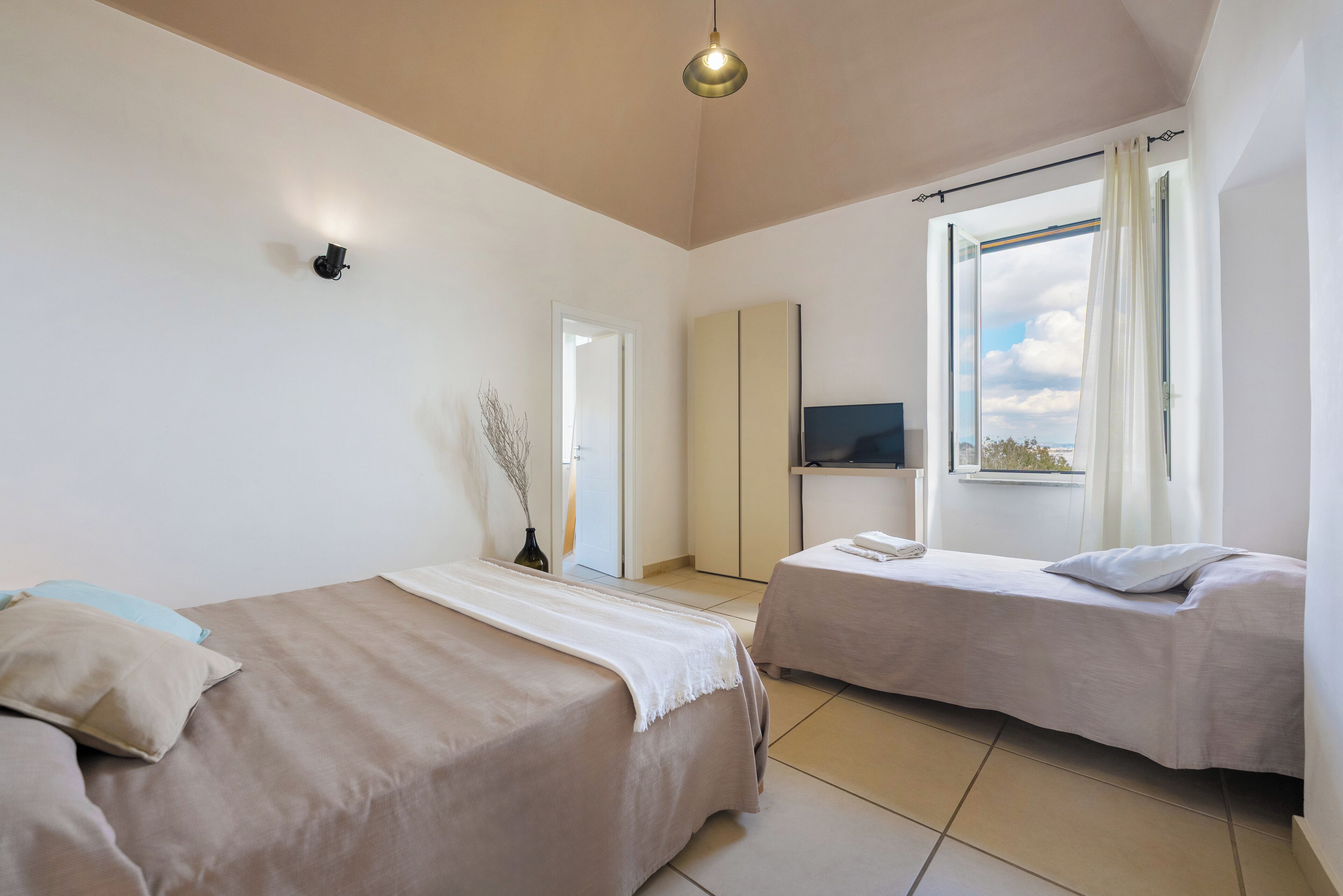 Una habitación de hotel con dos camas, un televisor, una ventana con cortinas y un jarrón con ramas.