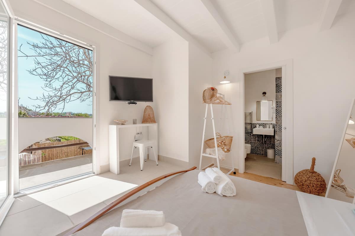 A modern, minimalist bathroom with a large window, a flat-screen TV, a white chair, a wooden bench, and a mirror.