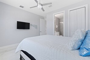 Un dormitorio con cama, ventilador de techo, televisor y baño visible a través de una puerta abierta.