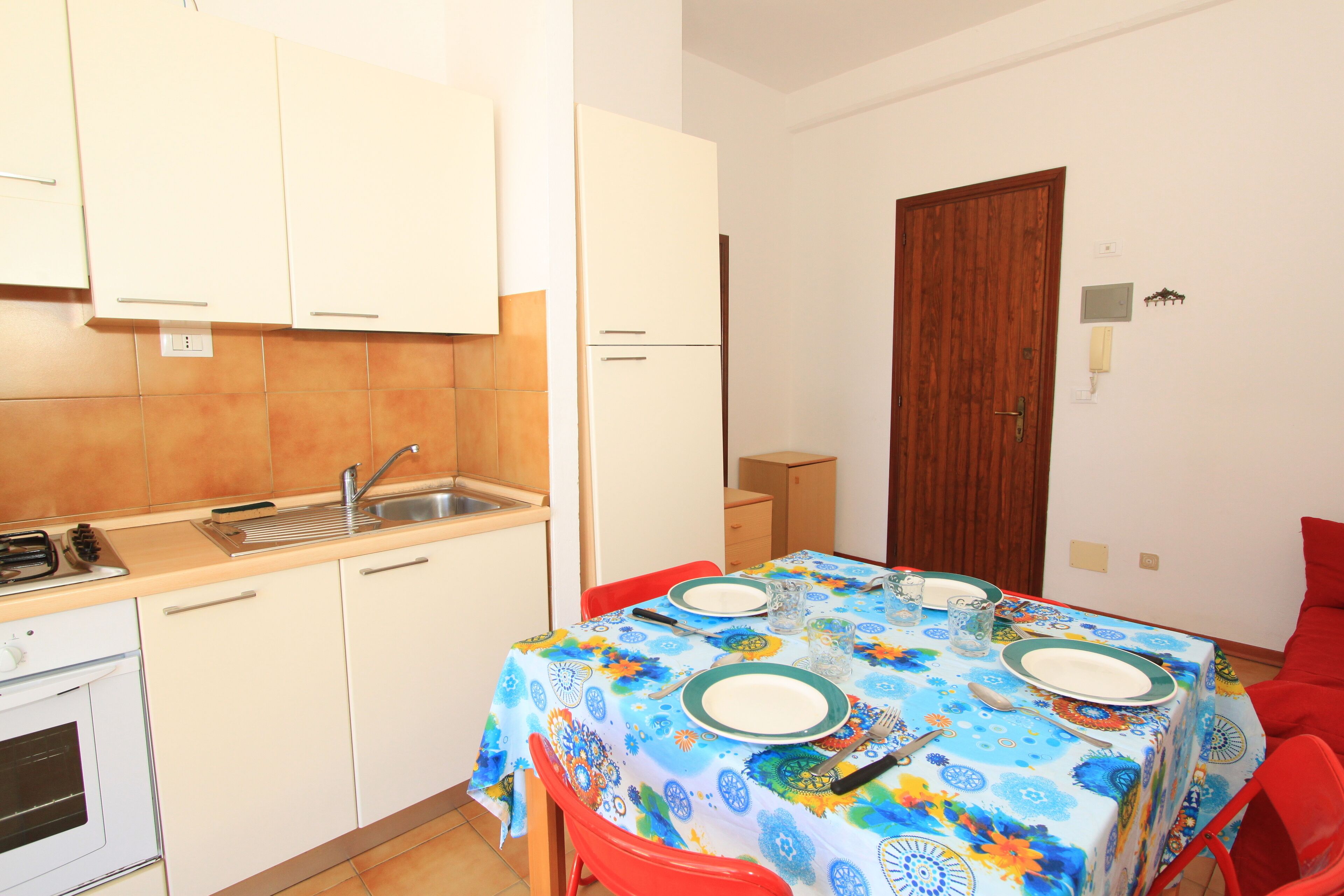 Una cocina compacta con armarios blancos, un fregadero y una estufa. Una mesa de comedor con mantel colorido y sillas rojas está instalada en la habitación.