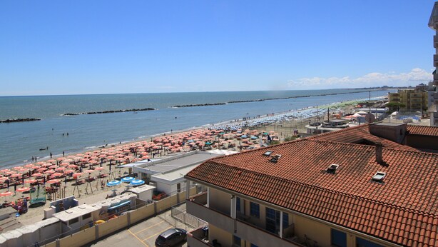 Un hôtel situé sur la côte, avec un toit recouvert de tuiles, une plage parsemée de parasols et une vue sur la mer.