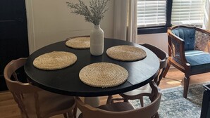 A dining area with a round table, four woven placemats, a vase with dried plants, and a framed picture of a cow on the wall. - Farmhouse - Centrally Located in Great Bend, Gas Firepit and Pet Friendly Yard (Great Bend)