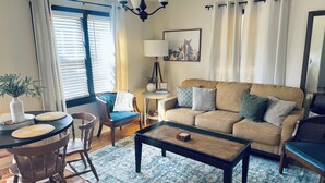 A living room with a beige sofa, a coffee table, a blue patterned rug, a dining area with a table and chairs, and a window with blinds. - Farmhouse - Centrally Located in Great Bend, Gas Firepit and Pet Friendly Yard (Great Bend)