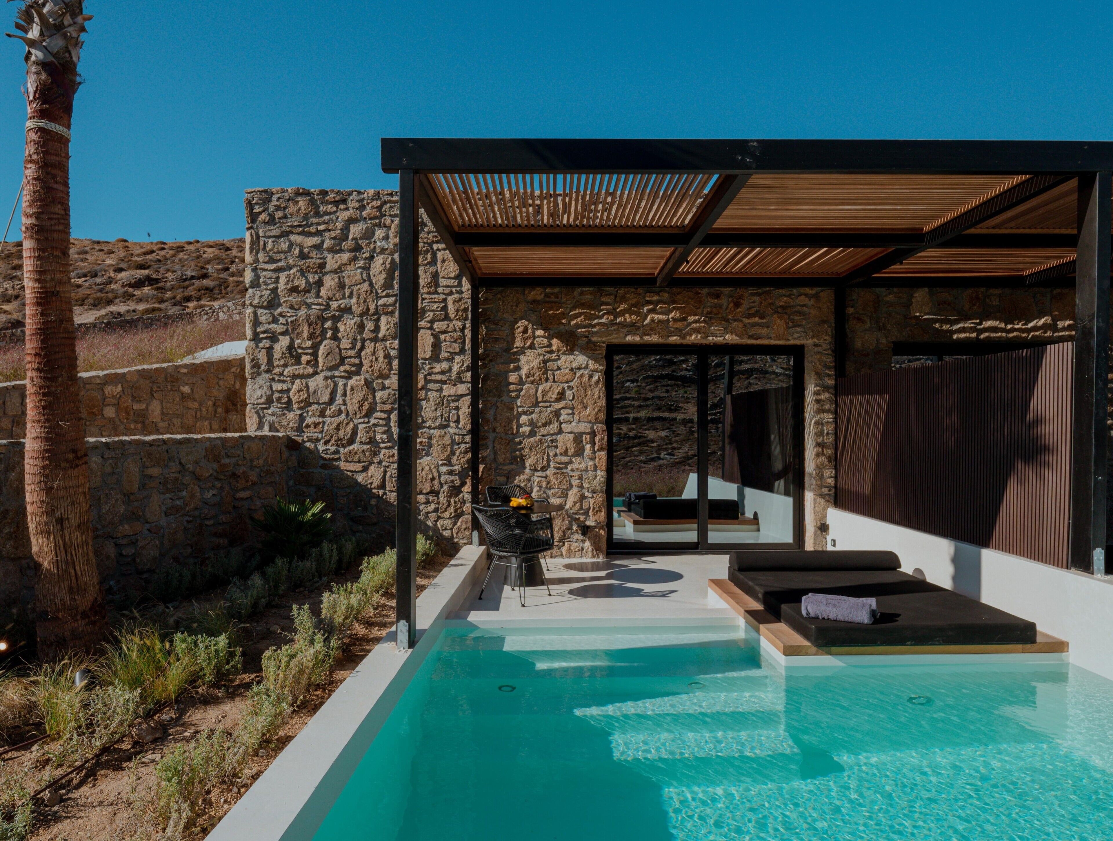 Área moderna da piscina ao ar livre, com uma parede de pedra, pérgula de madeira e um espaço de estar com mesa e cadeiras.