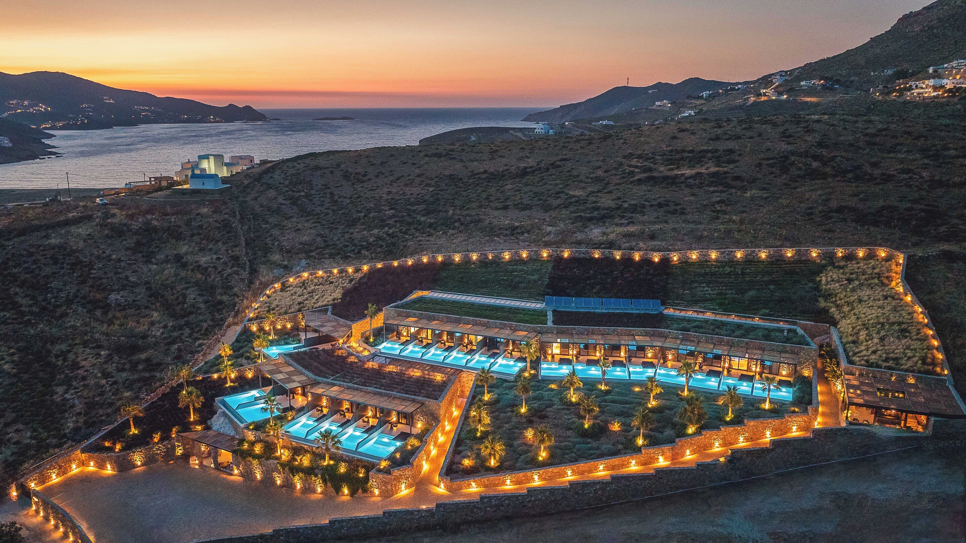 An aerial view of a resort with multiple swimming pools, a landscaped garden, and a view of the sea at sunset.