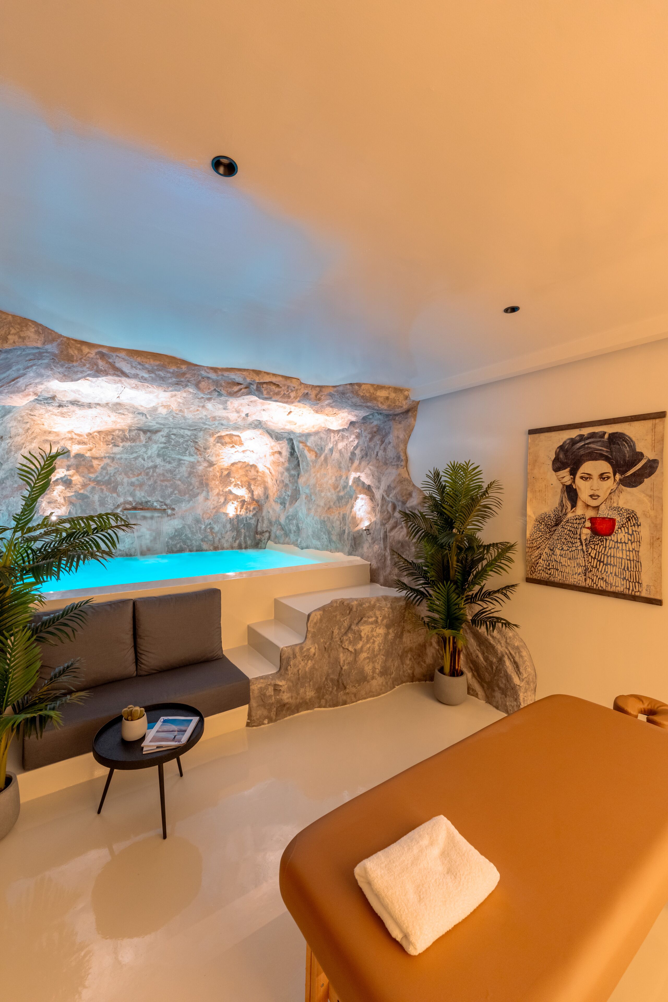 Un espacio de spa con una camilla de masaje, un rincón de descanso, una pared con textura rocosa y un elemento de agua.