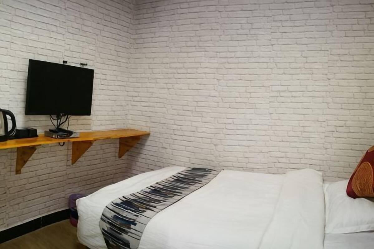 A room with a brick wall, a bed with a white and black patterned blanket, a TV on a wooden shelf, and a small table with a kettle.