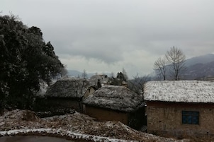 Un pueblo rural con casas de techo de paja tradicionales, un muro de piedra y montañas al fondo.